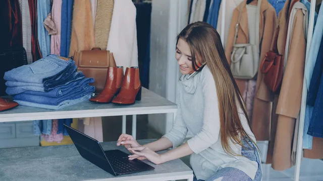 Young woman managing a fashion boutique, multitasking with phone and laptop, surrounded by clothing. How to read commerce reviews demand signals with a repeatable method