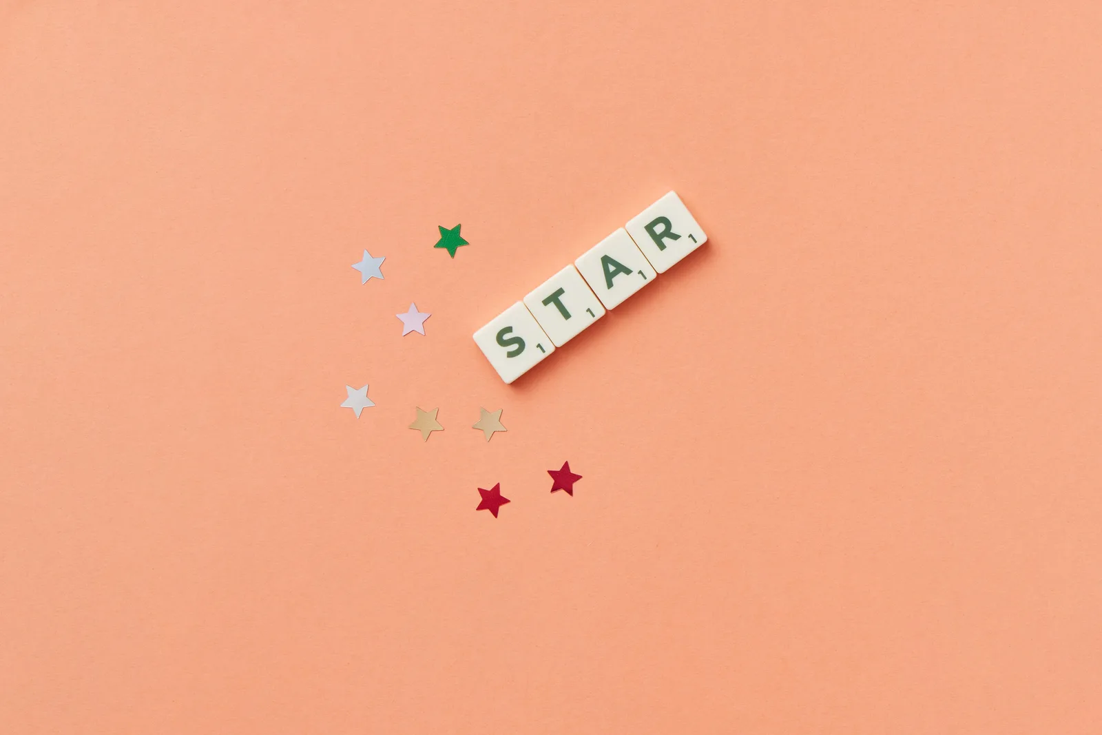 Flat lay image of Scrabble letters spelling 'STAR' on a peach background with colorful stars.