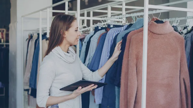 A woman browsing sweaters in a fashion retail store holding a tablet. When to build your commerce reviews foundations as a retailer: rules, data, costs and a 2027 checklist