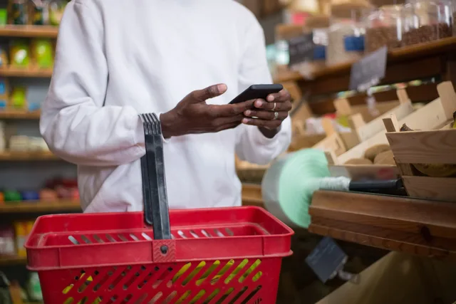 A man shops using a smartphone at a grocery store, holding a red basket filled with groceries. Commerce reviews launch review: is your process ready to launch before peak?