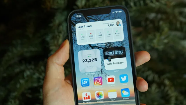 A hand holding a smartphone displaying various home screen widgets and app icons. Ecommerce UGC: planning a customer review pilot